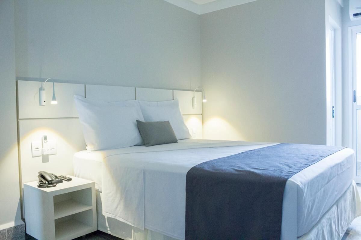 A modern hotel room with a neatly made bed, bedside lamps, a phone on the nightstand, and a window providing natural light.
