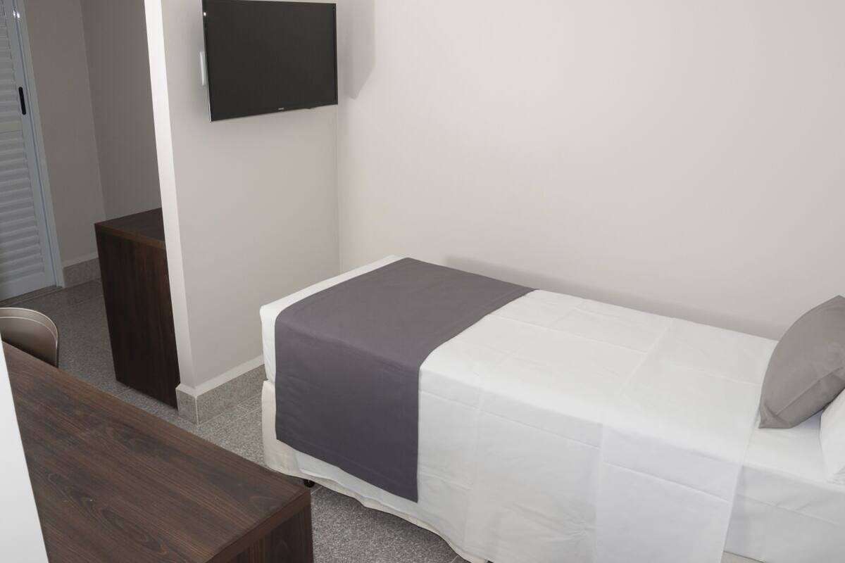 A small, neatly arranged bedroom with a single bed, a flat-screen TV mounted on the wall, and a wooden nightstand.