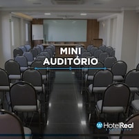 Uma sala de conferências com fileiras de cadeiras, um pódio e uma tela de projeção.