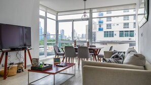 Una sala de estar moderna con un televisor grande, una mesa de comedor con sillas, un sofá y vistas a la ciudad a través de amplias ventanas.