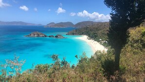 Un paisaje costero con playa arenosa, aguas turquesas cristalinas y colinas al fondo.