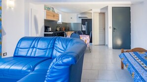 A living room with a blue sofa, a dining area with a table and chairs, a kitchen with appliances, and a door. - Apartment "Bleu Provence" (Les Salles-sur-Verdon)