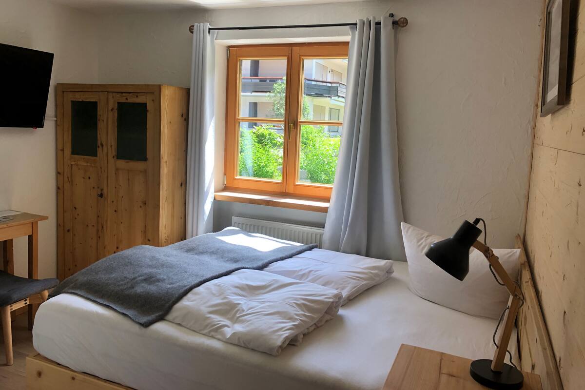 A bedroom with a bed, a desk, a chair, a window with curtains, and a flat-screen TV mounted on the wall.