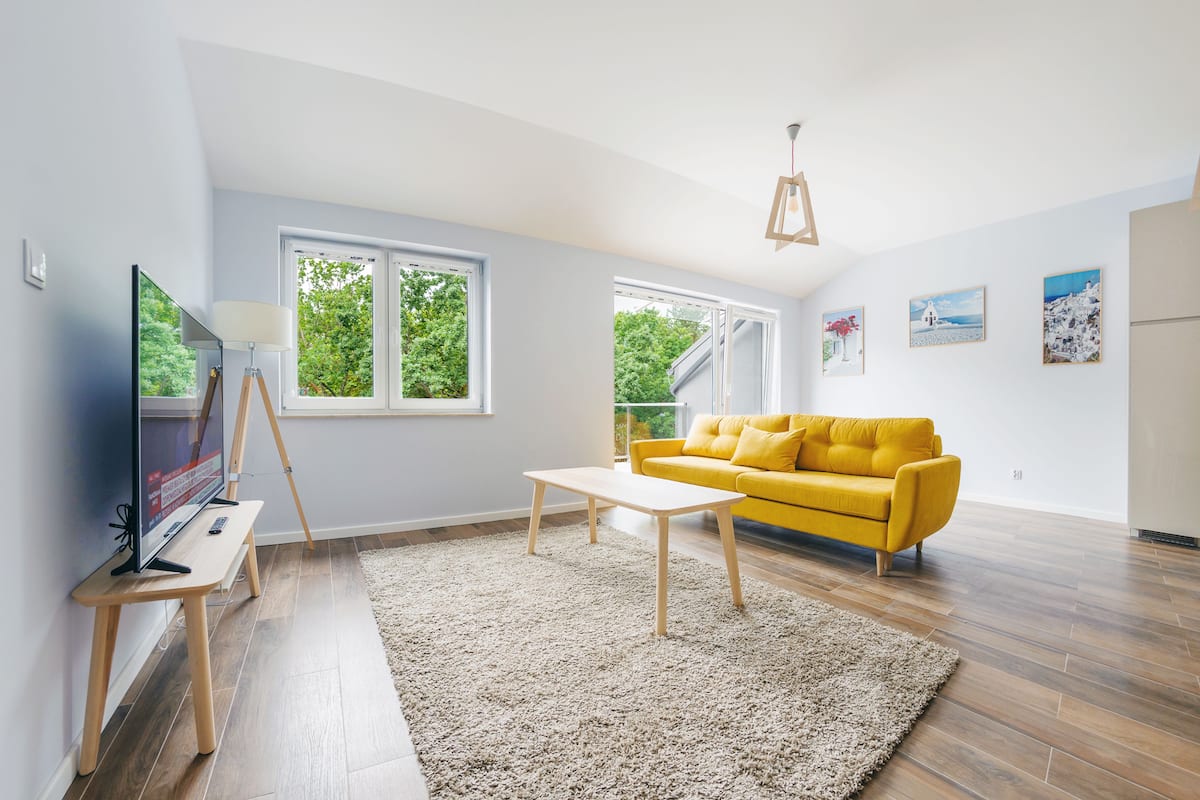 Un salon lumineux avec un canapé jaune, un téléviseur posé sur un meuble en bois et de grandes fenêtres donnant sur un paysage verdoyant.