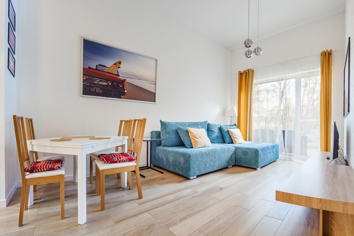 Un salon moderne avec une table à manger blanche, un canapé bleu et des chaises en bois. Un tableau représentant une voiture est accroché au mur, et une grande fenêtre est agrémentée de rideaux.