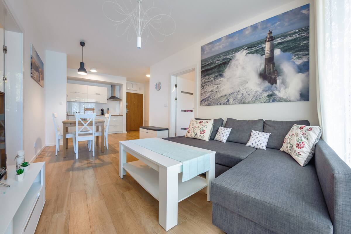 Un salon moderne avec un canapé gris, une table basse blanche et un tableau représentant un phare accroché au mur.
