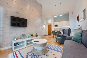 Una sala de estar moderna con un televisor de pantalla plana, un sofá gris, una mesa de centro de mimbre y una cocina al fondo.