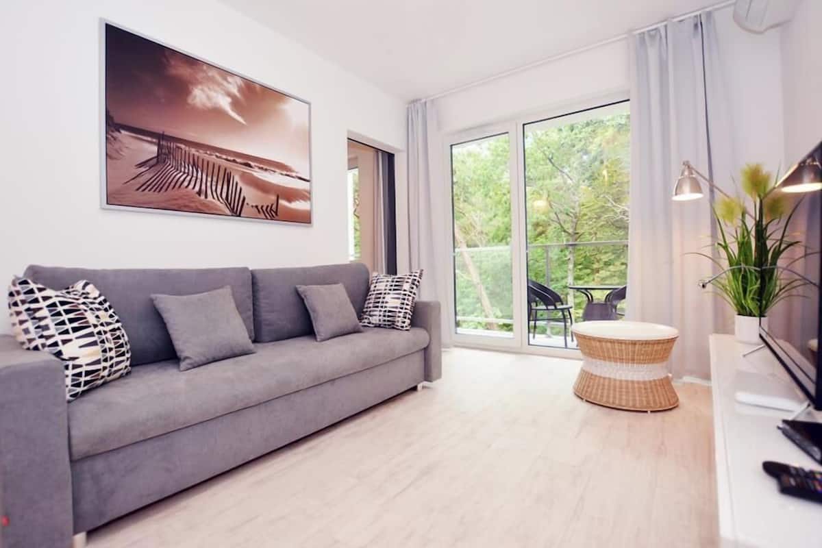 Un salon moderne avec un canapé gris, une grande fenêtre et une petite table d’appoint en osier.