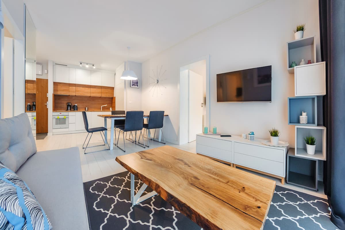 Un salon moderne avec une table basse en bois, un canapé gris et un coin repas comprenant des chaises, avec une cuisine en arrière-plan.