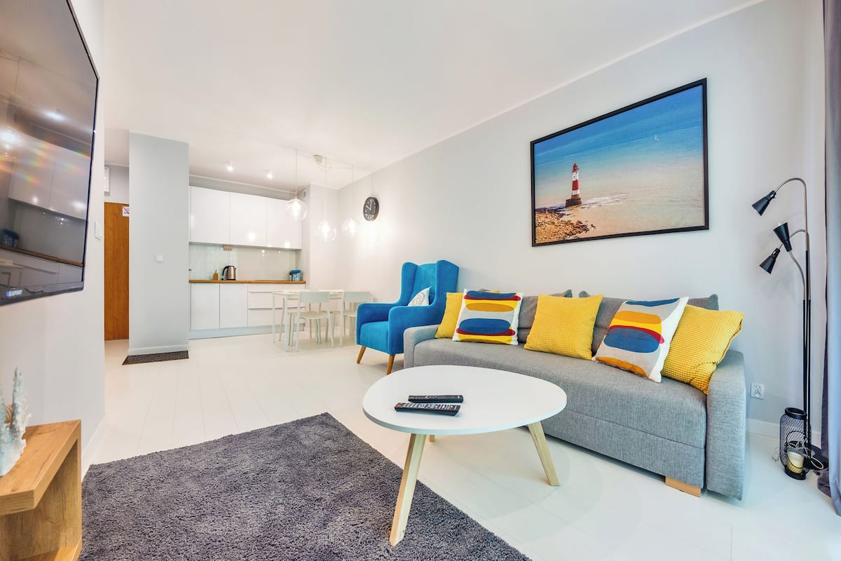 Un salon moderne avec un canapé gris, un fauteuil bleu et une table basse blanche. Un tableau représentant un phare est accroché au mur.