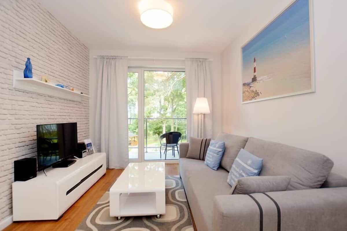 Un salon moderne avec un mur en briques blanches, un téléviseur à écran plat posé sur une console blanche, un canapé gris agrémenté de coussins bleus, une table basse blanche et un tableau représentant un phare.