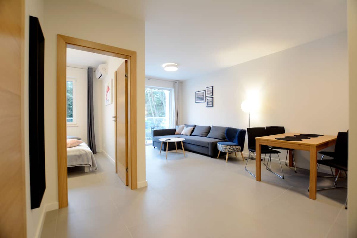 Un salon moderne avec un canapé, une table à manger et des chaises. On y trouve une fenêtre avec des rideaux, une porte donnant sur une autre pièce et un luminaire au plafond.