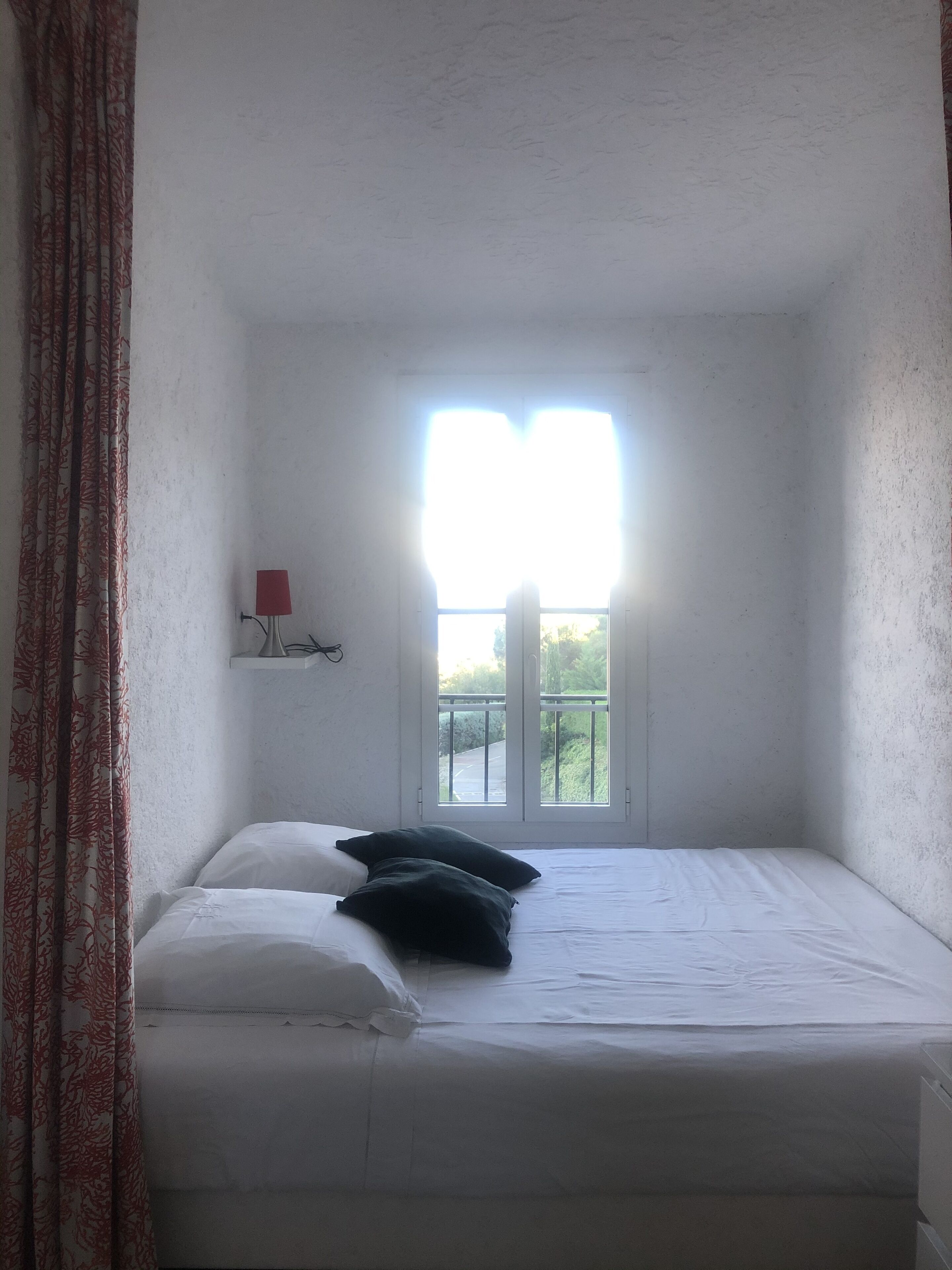 A small, single-bed room with a window, white walls, and a red lamp on a shelf.