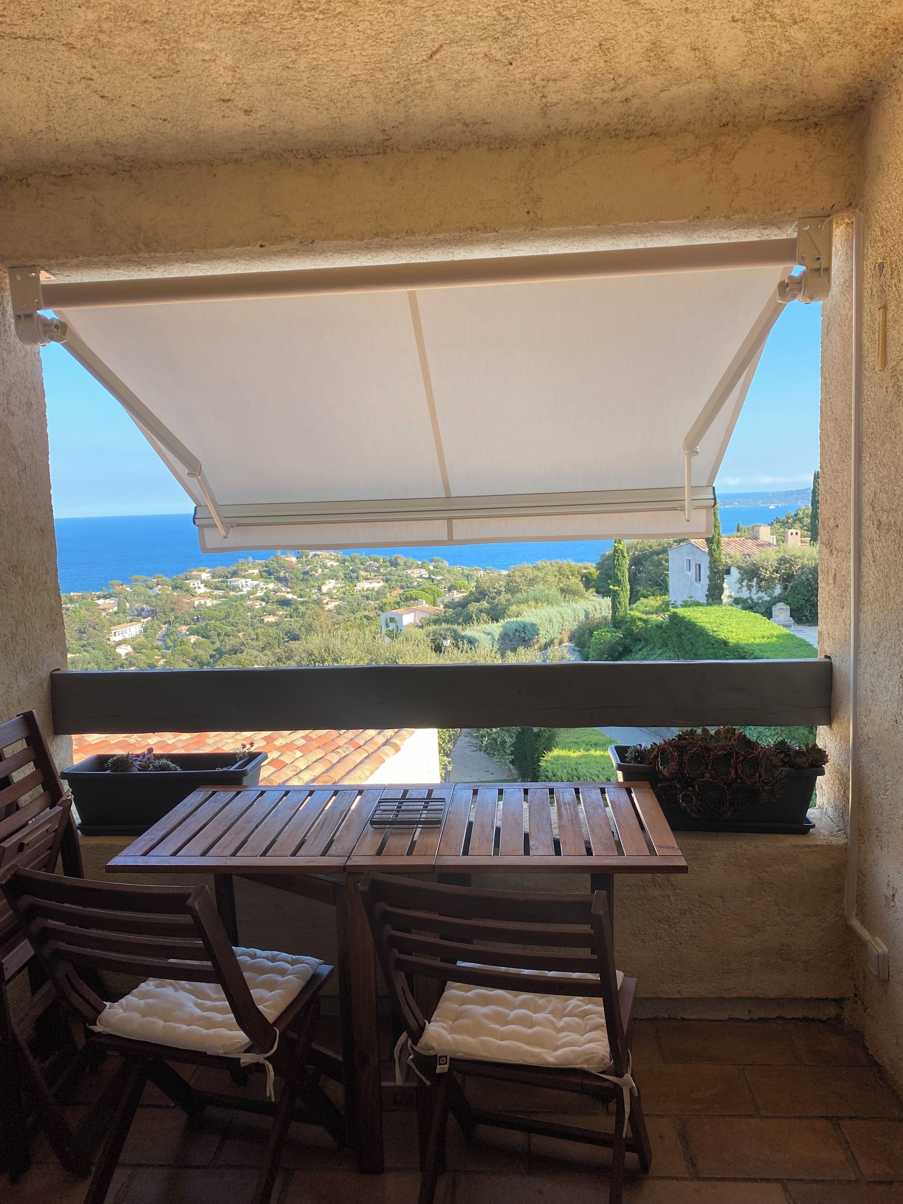 A balcony with a wooden table and chairs, a white awning, and a view of the sea and greenery.