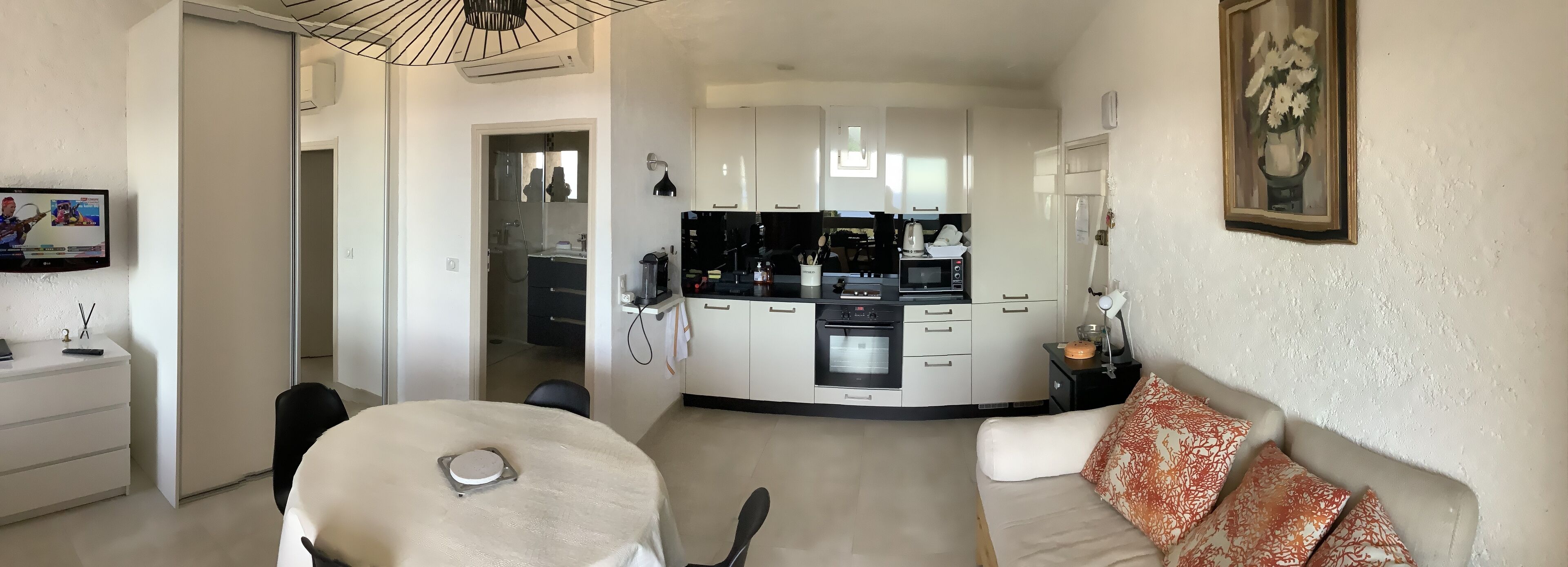 A modern kitchen with white cabinets, a black countertop, and a coffee machine. There is a dining area with a round table and chairs, and a sofa with patterned cushions.