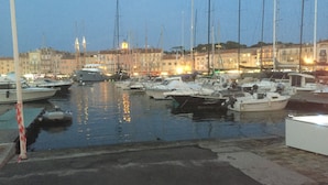 A marina with multiple boats docked, a waterfront promenade, and buildings in the background.