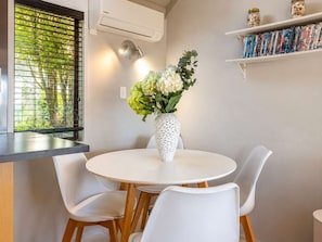Ein Essbereich mit einem runden Tisch, weißen Stühlen, einer Vase mit Blumen, einem Fenster mit Jalousien, einer Klimaanlage und einem Bücherregal mit Büchern.