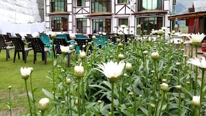 Un edificio con un estilo arquitectónico tradicional, con un gran ventanal y un techo verde. Hay zonas de estar al aire libre con sillas y mesas de plástico, y un jardín con flores blancas en primer plano.