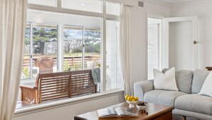 A living room with a large window, a wooden bench, a grey sofa, and a table with a bowl of fruit and an open book. - Pippi House - A Quintessential Beach House Opposite Pippi Beach - Holiday Yamba (YAMBA)