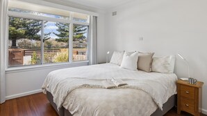 A bedroom with a large window, a bed with multiple pillows, a bedside table with a lamp, and a small wooden nightstand with drawers. - Pippi House - A Quintessential Beach House Opposite Pippi Beach - Holiday Yamba (YAMBA)