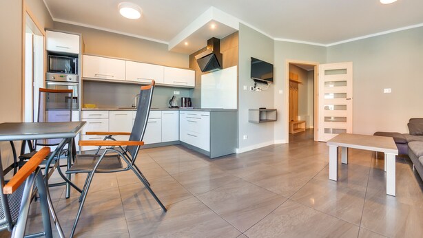 A modern kitchen with white cabinets, a built-in oven, and a dining area with a table and chairs. - Apartamenty Sun & Snow Osiedle Wybrzeże (Ustronie Morskie)