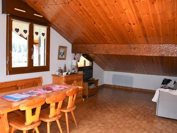 Ein gemütliches Dachzimmer mit Holzbalken, einem Esstisch mit Stühlen, einem Fernseher und einer Couch.