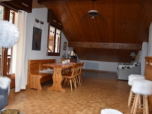 Ein gemütlicher Essbereich mit Holztisch und Bänken, im Hintergrund eine Küche und eine Decke mit freiliegenden Balken.