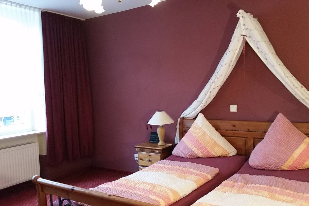 A bedroom with a bed under a canopy, a nightstand with a lamp, and a window with curtains.