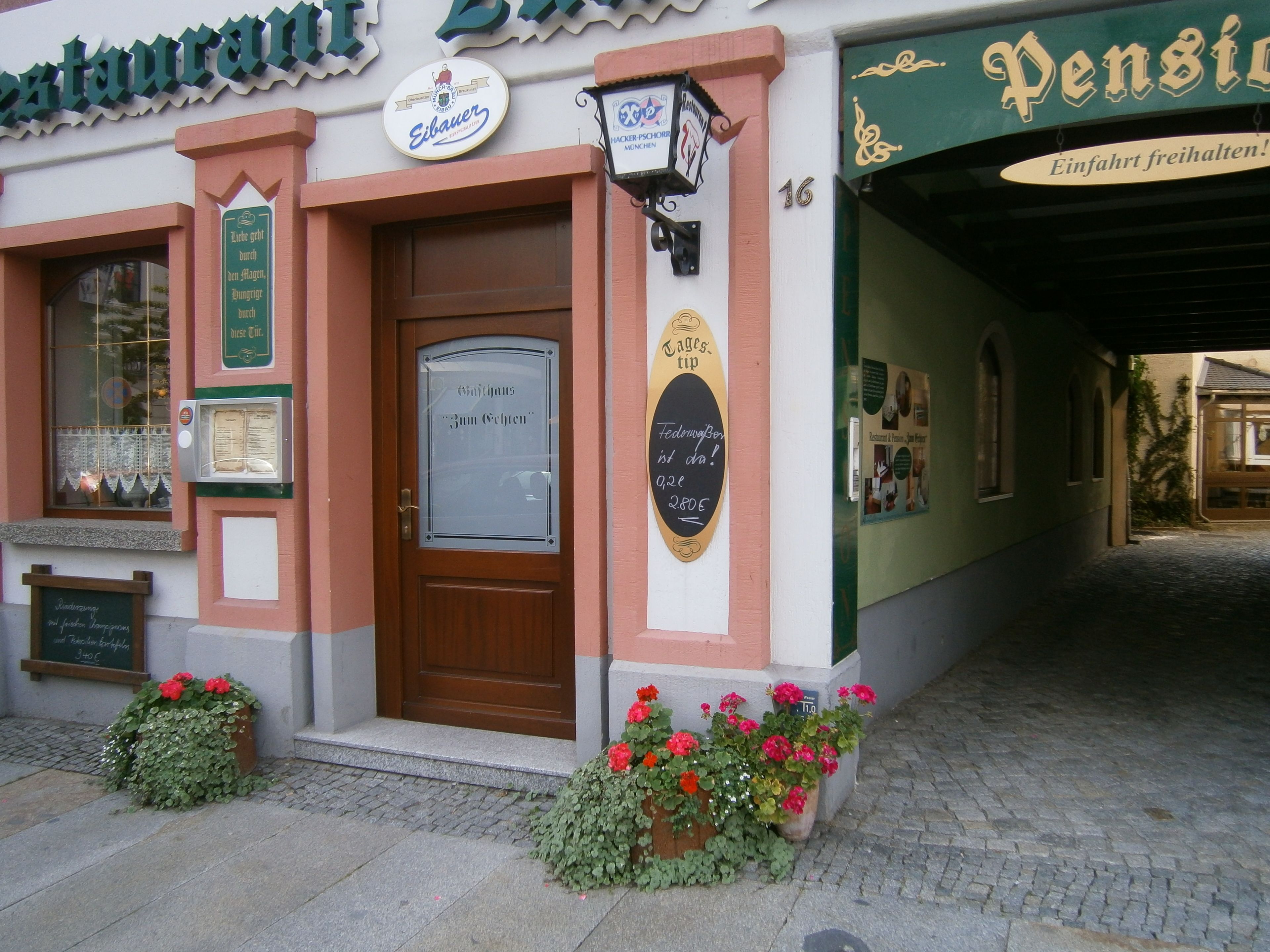 A traditional European building with a wooden door, signs for 'Eibauer' and 'Pension', and flower boxes on the ground.