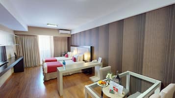 Habitación de hotel con dos camas, un televisor, un escritorio con una silla, una pequeña mesa de comedor con una botella de agua y un plato de fruta, y un aire acondicionado instalado en la pared.