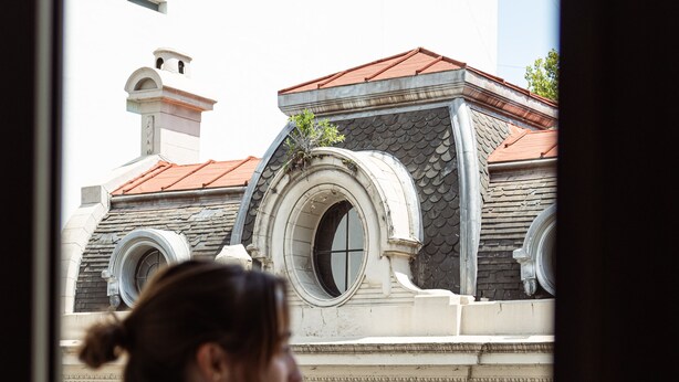 Una persona de pie junto a una ventana con vistas a un edificio de arquitectura ornamentada y techo rojo.