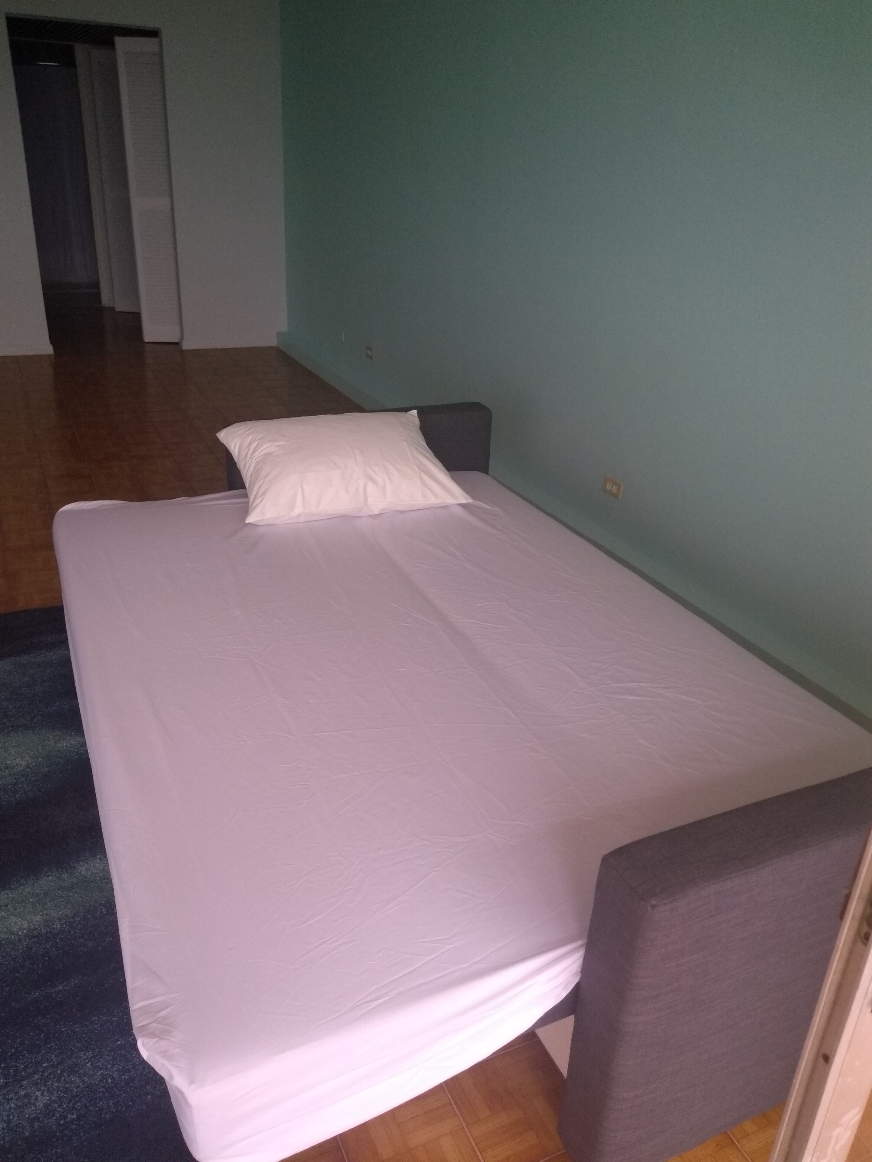 Un lit simple avec un matelas et un oreiller blancs, dans une pièce aux murs de couleur turquoise et au plancher en bois.