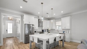 Cocina moderna con armarios blancos, una isla central con taburetes de bar, electrodomésticos de acero inoxidable e iluminación colgante.