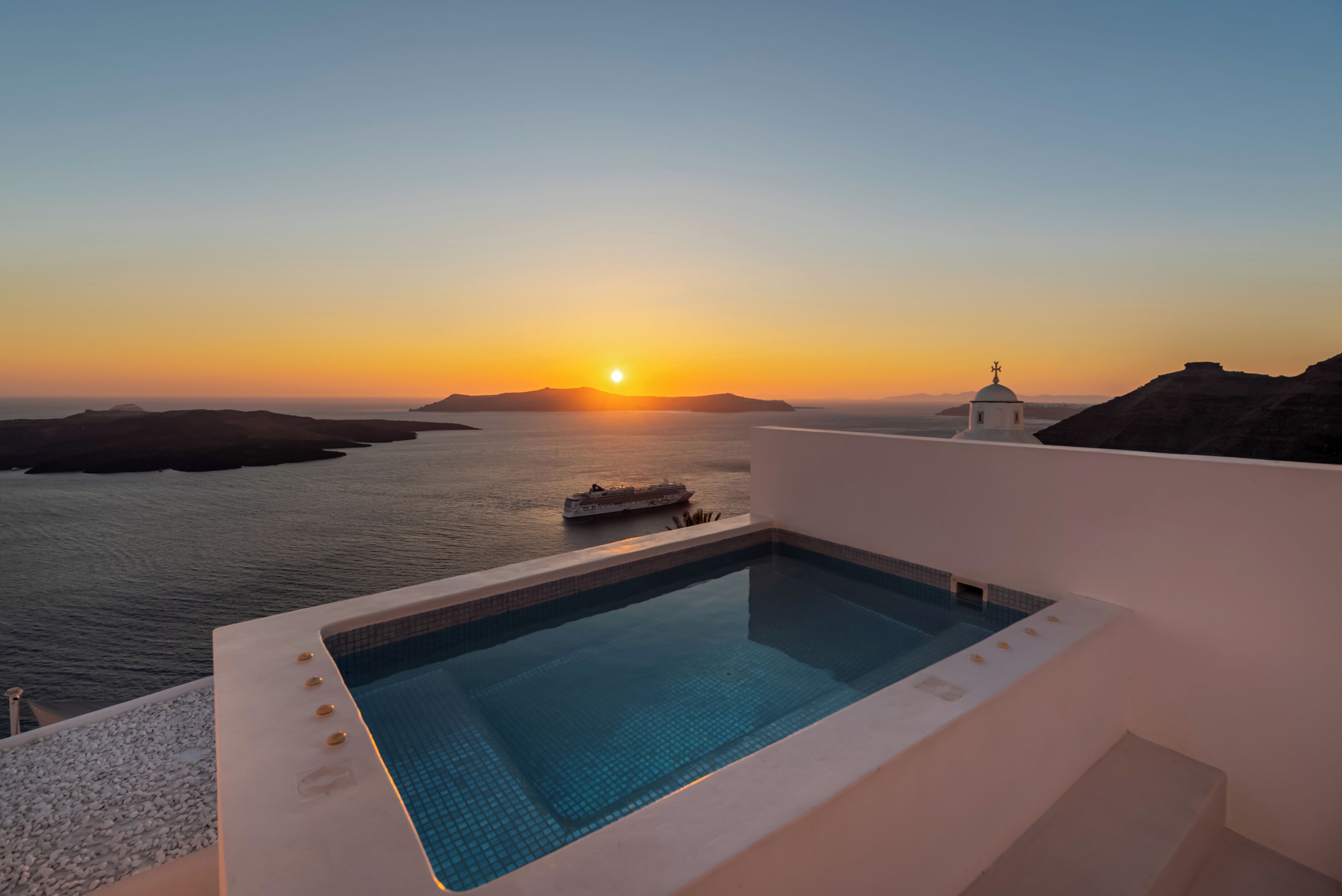 Ein Pool mit Blick auf den Sonnenuntergang über dem Meer und ein kleines Gebäude mit einem Kreuz auf dem Dach.