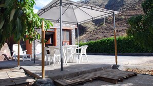 A covered outdoor seating area with white plastic chairs and tables, a wooden deck, and a mountain backdrop.
