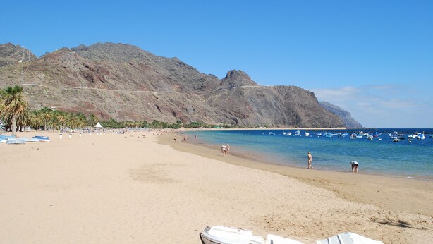 A beach with a rocky mountain in the background, a clear sky, and several boats in the water.