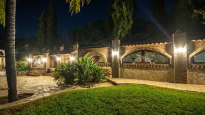 A well-lit outdoor area with stone pathways, palm trees, and a decorative wall with arches. - Cubo's Mijas Finca Huerta Rica Sabana / shared pool / views (Mijas Pueblo)