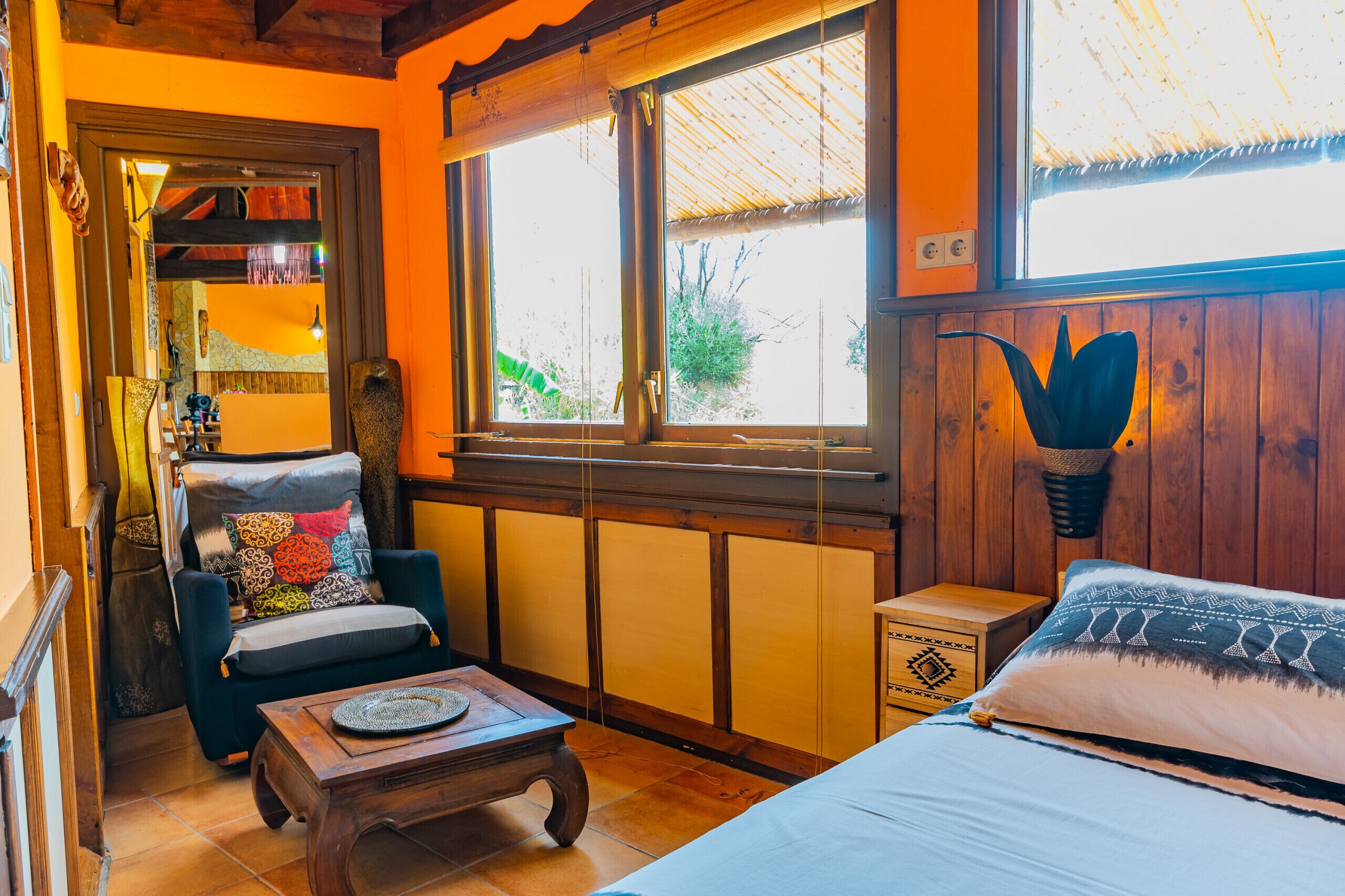 Una habitación acogedora con una cama, una silla con cojines coloridos, una mesa de madera y una ventana con vistas a zonas verdes.