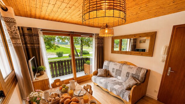 A cozy room with a wooden ceiling, a sofa, a dining table with breakfast items, and a balcony with a view of greenery.