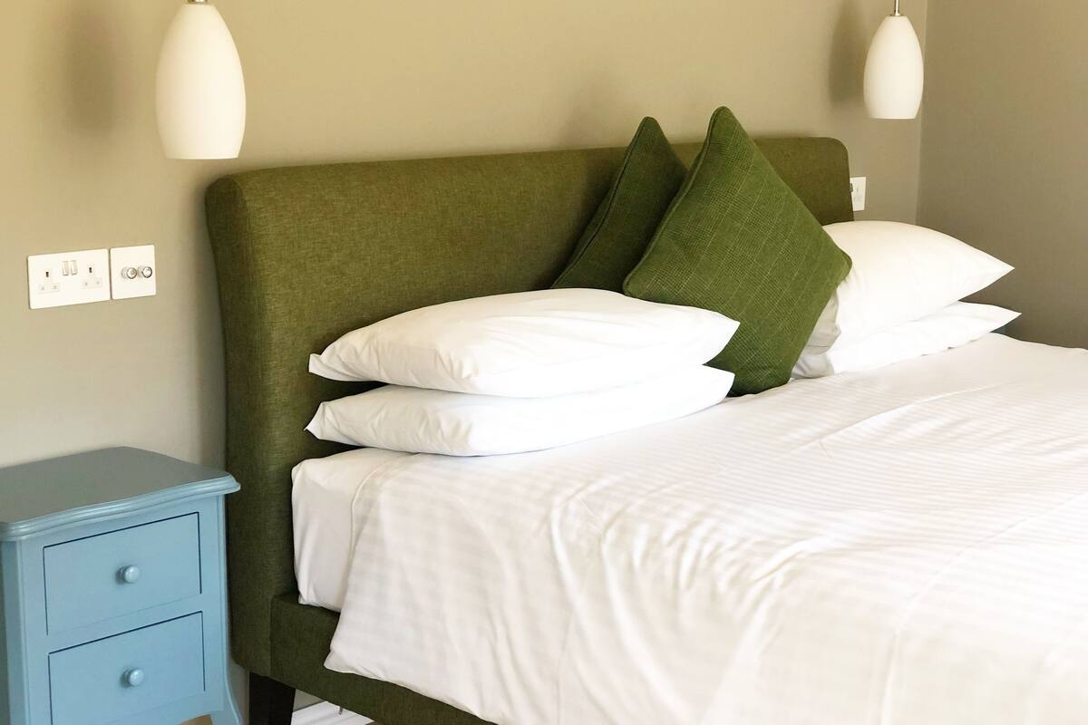 A neatly made bed with white linens, a green upholstered headboard, and matching green pillows. There is a small blue nightstand with drawers to the left.