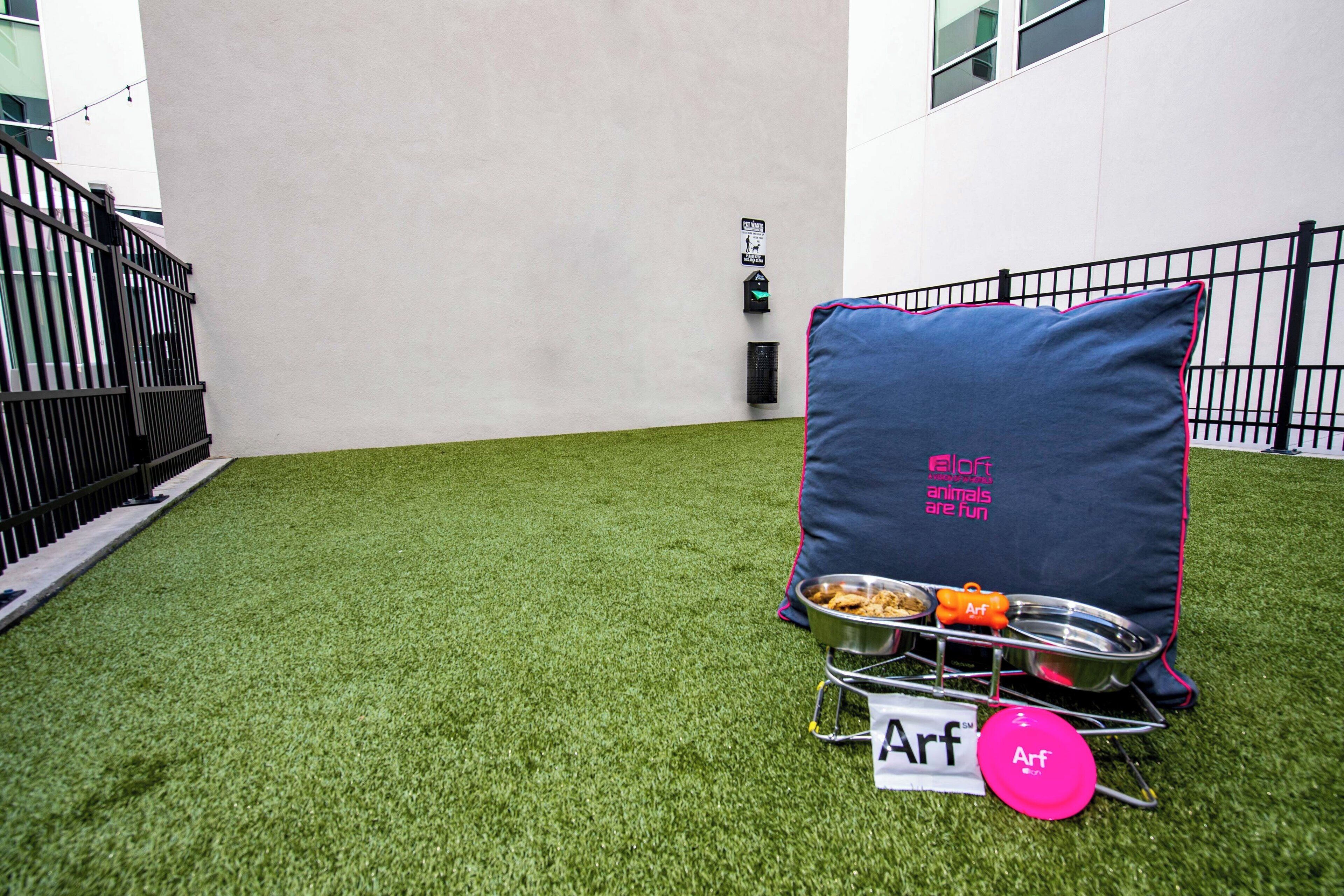 Un petit espace extérieur avec du gazon synthétique, une chaise en métal avec un coussin, un plateau en métal pour la nourriture et un objet rose portant l’inscription « Arf ».