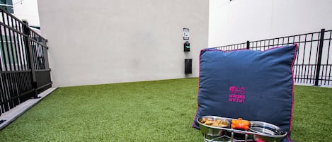 Un pequeño espacio al aire libre con césped artificial, una silla de metal con cojín, una bandeja metálica para alimentos y un objeto rosa con la etiqueta 'Arf'.