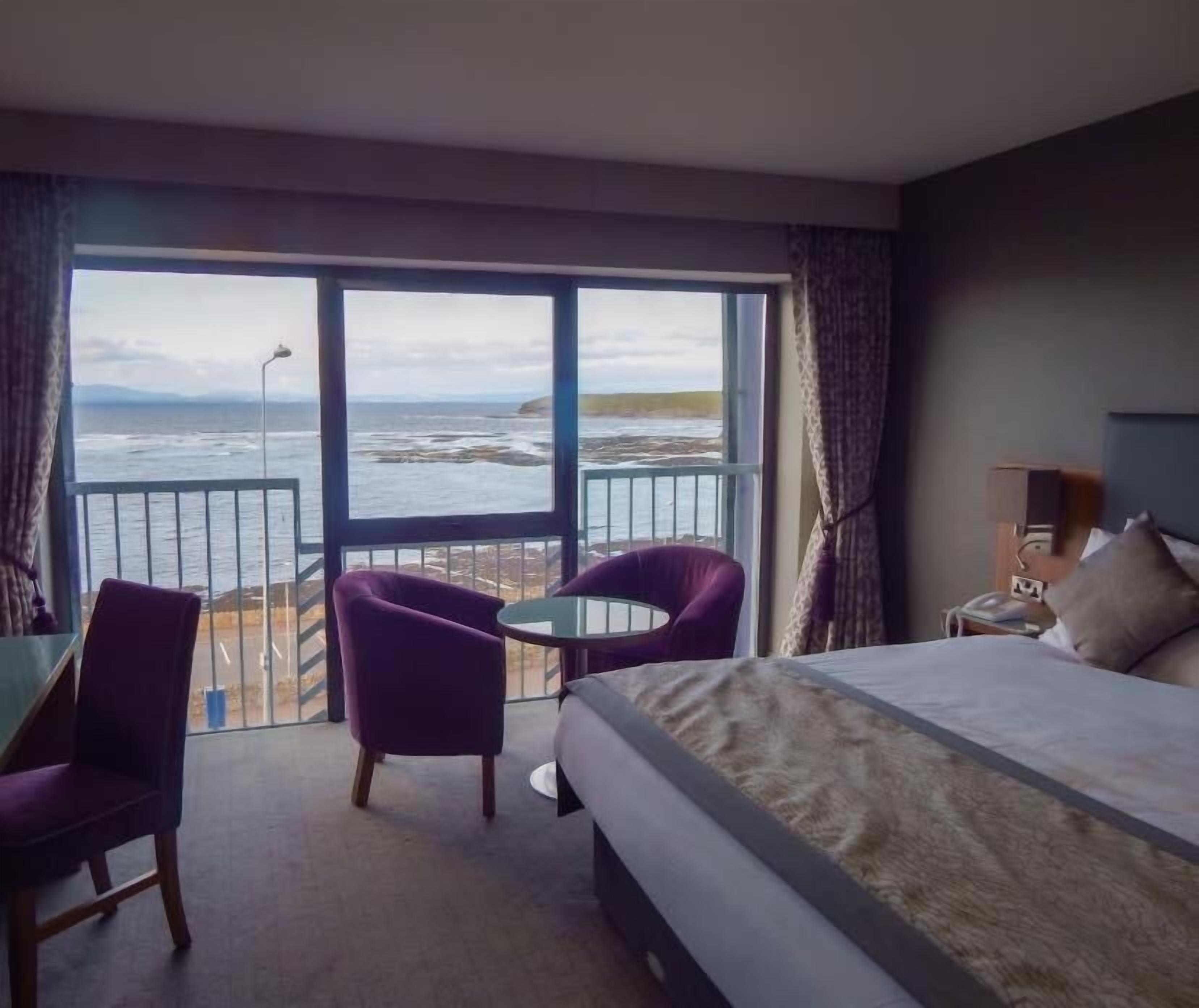 A hotel room with a large bed, a small table, two purple chairs, and a balcony with a view of the sea.