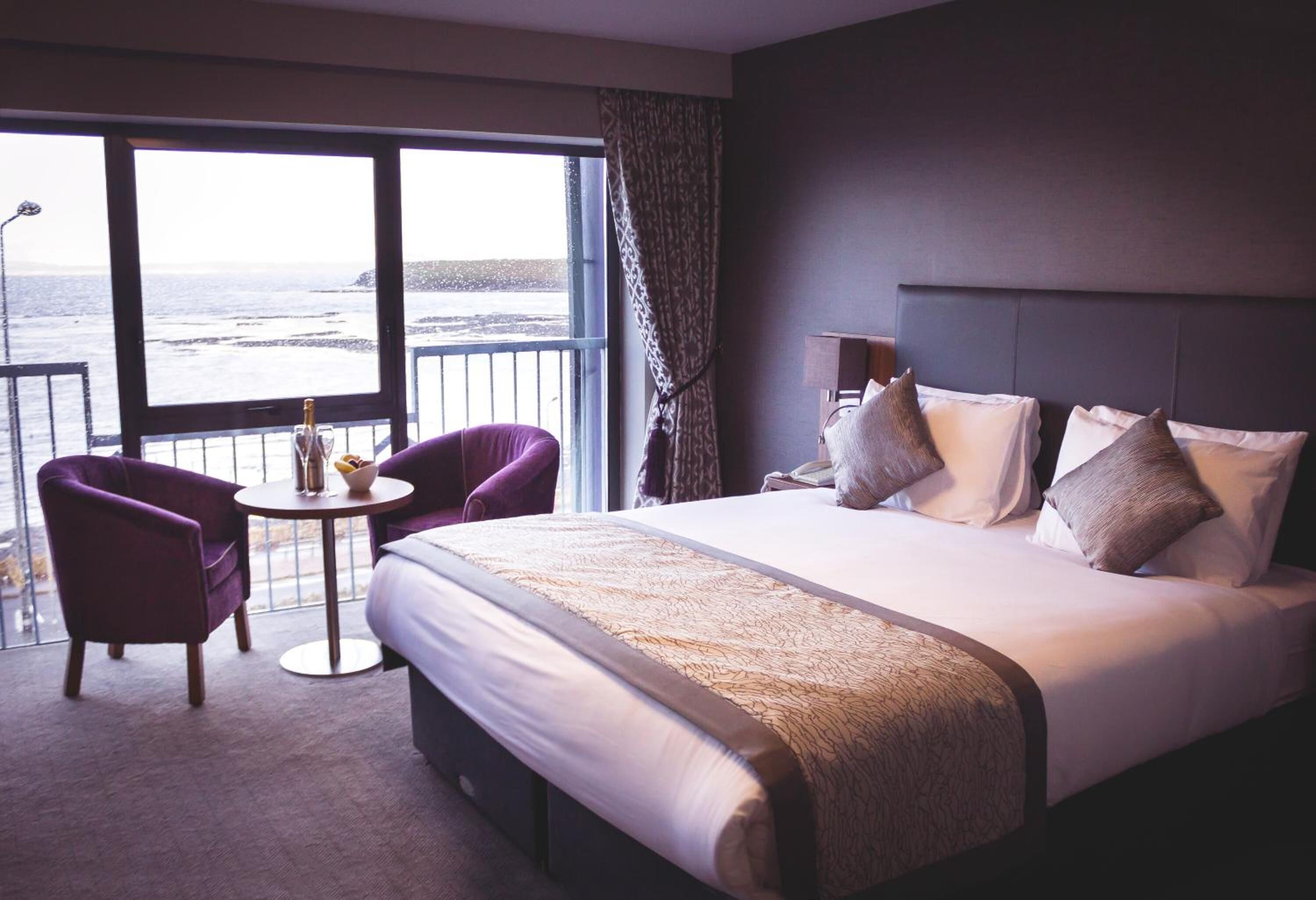 Quarto de hotel com uma cama grande, uma mesa redonda pequena, duas poltronas roxas e vista para o oceano através de grandes janelas.