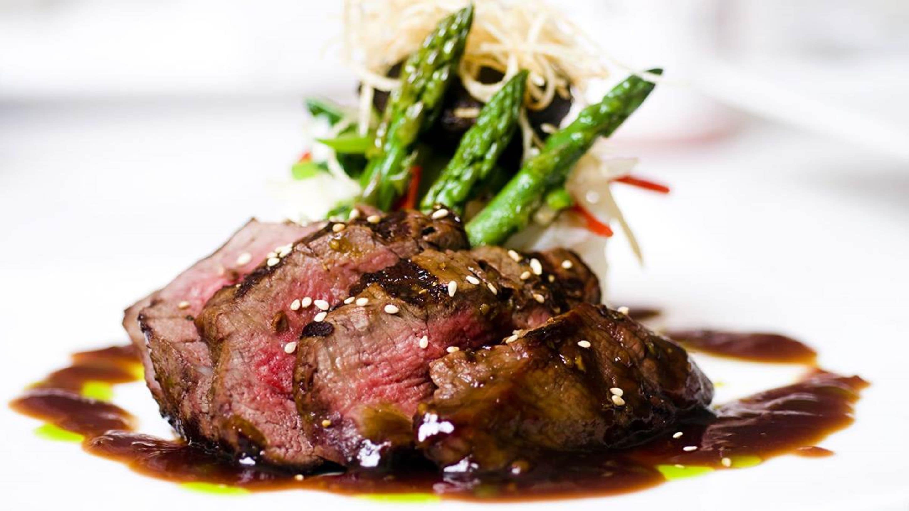 A steak with sesame seeds and a sauce, asparagus, and a side of noodles on a white plate.