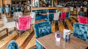 A restaurant with velvet chairs in blue and pink, wooden tables, and a bar area with a wooden counter and shelves of bottles.