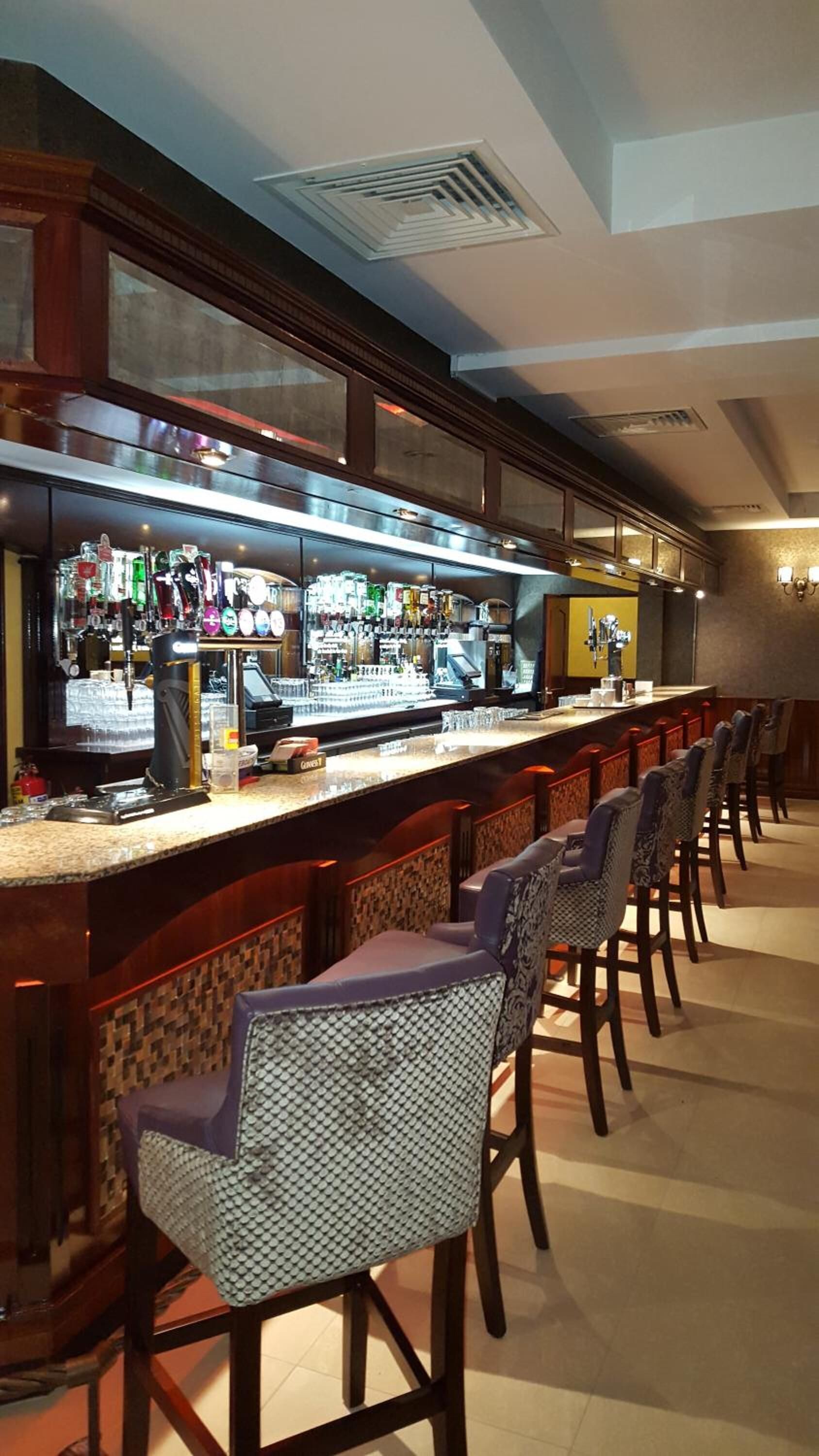 Eine Bar mit einer langen Theke, mehreren Zapfhähnen und einer Auswahl an Flaschen. Mehrere gepolsterte Stühle mit einem Muster sind ebenfalls vorhanden.