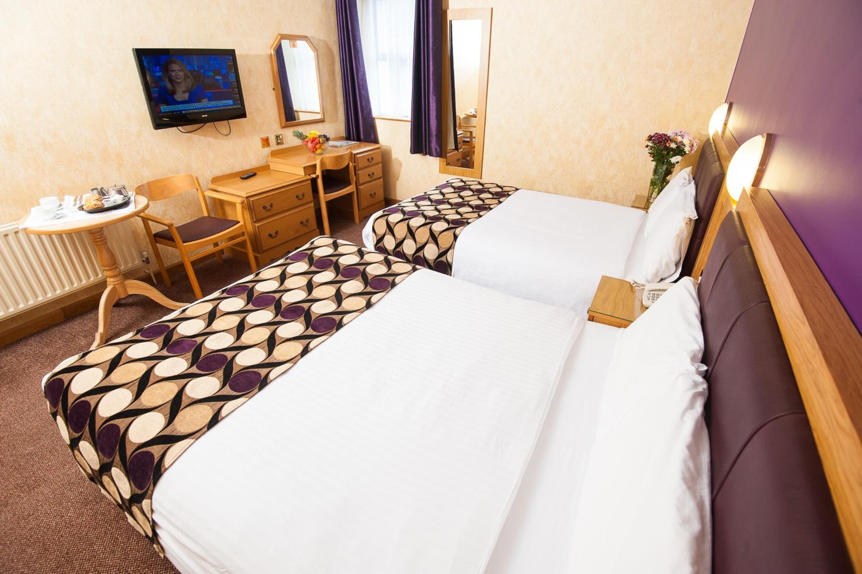 A hotel room with two beds, a desk with a chair, a television, and a small table with a vase of flowers.