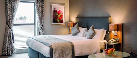 A hotel room with a large bed, two bedside lamps, a bedside telephone, a small round table with a fruit bowl, and a window with curtains.