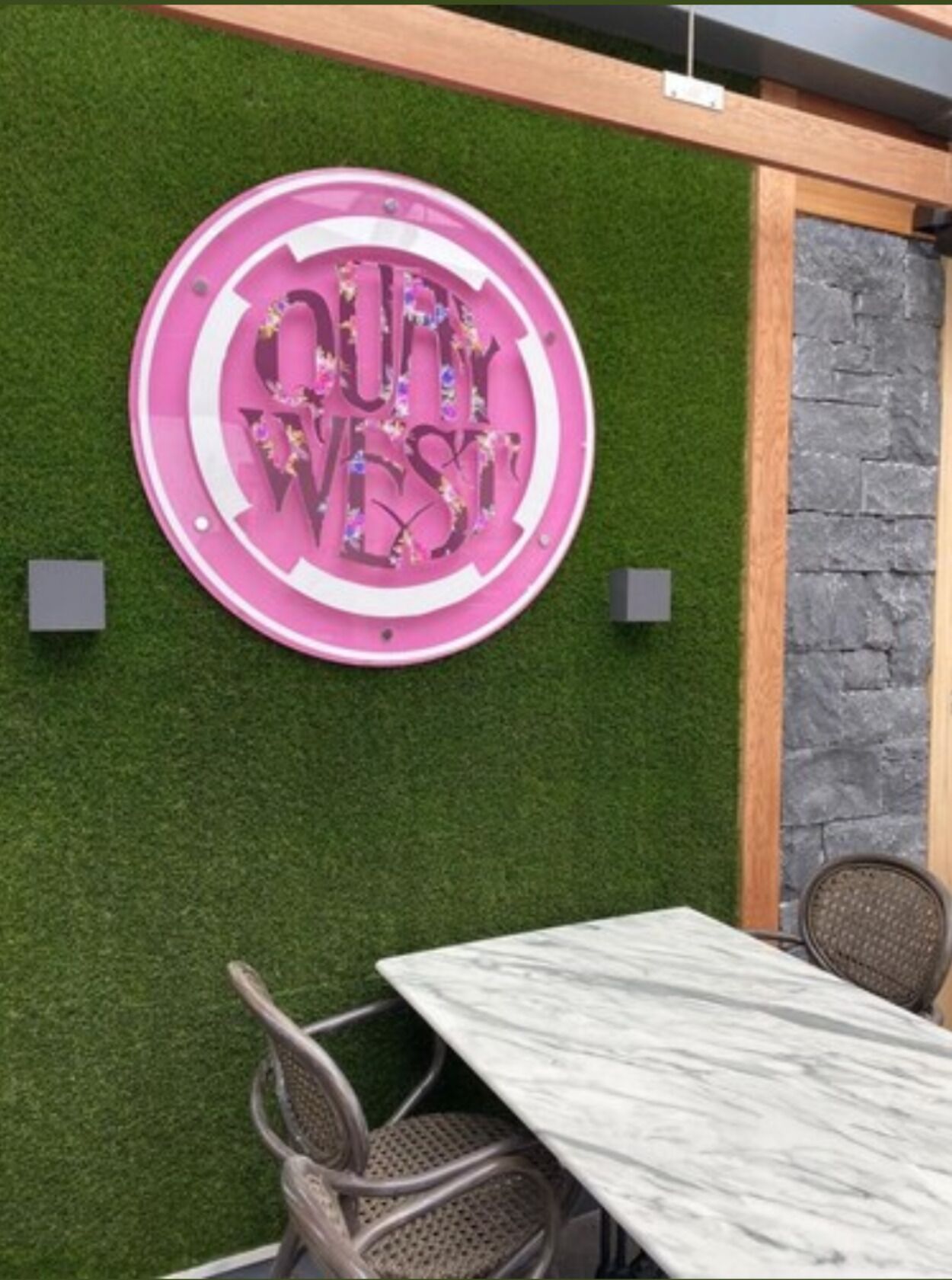 A circular sign with the text "OUT WEST" on a green artificial grass background, a white marble table, and wicker chairs.
